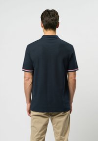 Tummansininen polo-paita lyhyillä hihoilla, jossa on ribbireunus kauluksessa ja punavalkoiset raidat hihoissa. Sileä kangas, klassinen istuvuus.