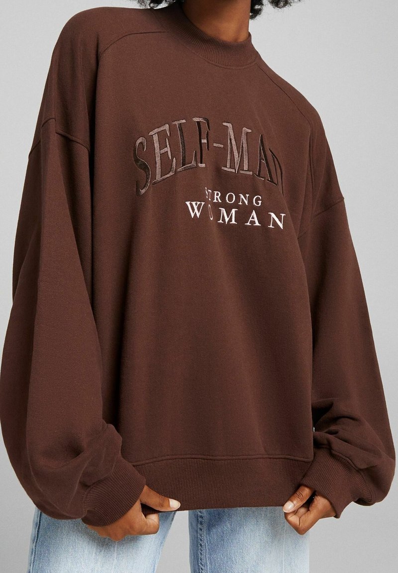 Bruine oversized sweatshirt met geribbelde halslijn en manchetten, met de tekst "SELF-MADE STRONG WOMAN" in contrasterende kleuren.