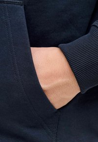 Tumedat sinine sviiter, close-up, rõhutades tekstuurset materjali, soonikkoes varrukaid ja kätt, mis toetub küljetaskusse.