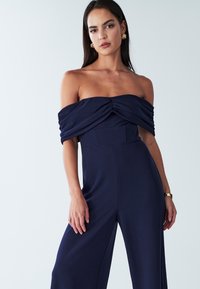 Marineblauwe off-the-shoulder jumpsuit met een gathered lijfje, brede benen en een gladde stof. Minimalistisch ontwerp, aangesloten taille en een zachte textuur.