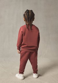 Sweatshirt rouge et joggers assortis, tous deux en tissu doux, avec des poignets côtelés et une coupe décontractée. Des baskets blanches complètent la tenue.