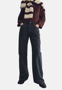 Schwarze Jeans mit weitem Bein, kombiniert mit einer braunen Wildlederjacke in Kurzform und einem beigen, gestreiften Schal. Strukturierte Stoffe, legere Passform.