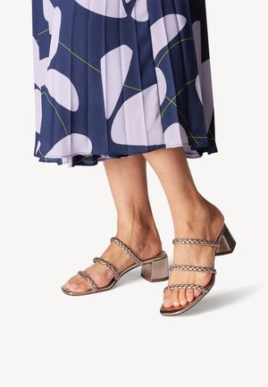 Strappy block-heeled sandals in metallic bronze; three braided straps, open toe design, and a textured heel. Worn with a patterned skirt.