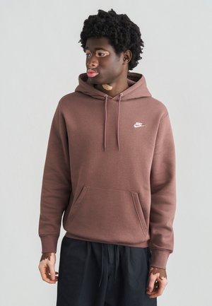 Sudadera con capucha marrón de Nike con bolsillo frontal, cordones ajustables y un pequeño logo blanco en el pecho. Modelo con pantalones negros.
