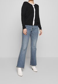 Svart stickad cardigan med kontrasterande vit knappfält, kombinerad med ljusblå utsvängda jeans och vita klackskor.