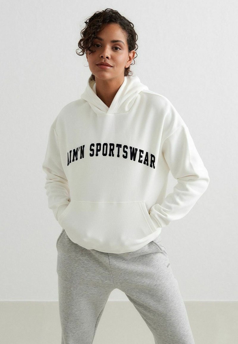 Sudadera blanca con "AIM'N SPORTSWEAR" impreso en negro. Presenta un bolsillo canguro y capucha con cordón. Combinada con pantalones de chándal gris claro.