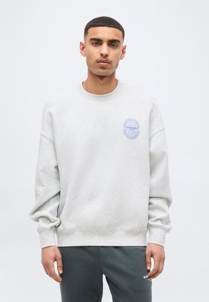 Νέος άνδρας με κοντά μαλλιά, φοράει γκρι ανοιχτό φούτερ με μικρό μπλε λογότυπο υδρόγειου και σκούρο γκρι παντελόνι, στέκεται μπροστά σε λευκό φόντο.