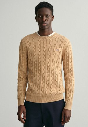 Beige kabelgebreide trui met een gestructureerd ontwerp, ronde halslijn en geribbelde manchetten en zoom, met een klein logo op de borst.