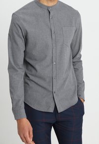 Camicia grigia a maniche lunghe con colletto rotondo, fronte con bottoni e taschino. Realizzata in tessuto testurizzato, presenta un design aderente.