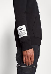 Zijaanzicht van een persoon die een zwarte hoodie draagt met een Alpha Industries-logo patch op de mouw en een zwarte broek met een ritszak.