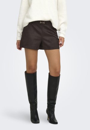 Kvinde iført mørkebrune lædershorts, sorte knælange stiletstøvler og en hvid, blød sweater, der står foran en ensfarvet baggrund.