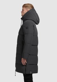 Podšívaná čierna parka s veľkým kapucňom, ktorá obsahuje zips, bočné vrecká a okrúhlu nášivku s logom na rukáve. Hladká, štruktúrovaná tkanina.