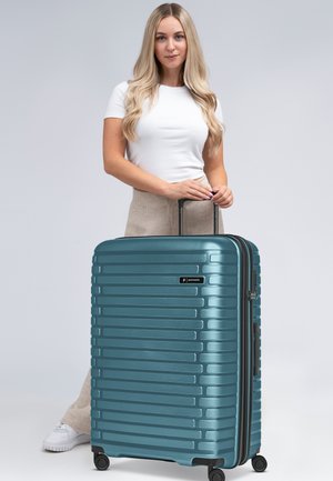 Maleta rígida en color verde azulado con rayas horizontales, asa retráctil, cuatro ruedas y una etiqueta de marca. Acabado de plástico suave y duradero.