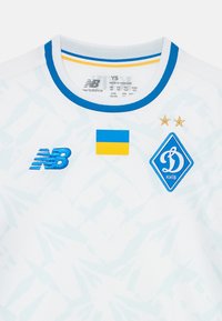 Bela kratka športna majica s kratkimi rokavi z modrimi poudarki, ki vsebuje modro "NB" logo, ukrajinsko zastavo in diamantno obliko emblema Kijeva z zvezdami.
