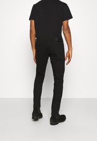 Man som bär svarta slim-fit jeans, svart t-shirt och svarta ankelstövlar, stående mot en enkel vit bakgrund med ryggen vänd mot kameran.