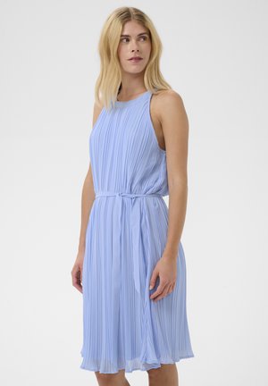 Kobieta w bezrękawnikowej, jasnoniebieskiej plisowanej sukience z paskiem w talii, stojąca i patrząca w bok na jednolitym tle.
