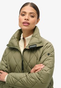 Veste matelassée en olive avec un col montant, fermeture éclair et logo sur le col. Tissu texturé avec une coupe décontractée.
