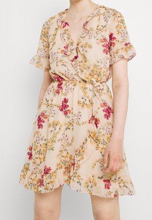 Frau trägt ein beiges, geblümtes Wickelkleid mit gerafften Ärmeln und Saum, mit gelben und roten Blumenmustern.