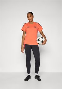 Oranje sportshirt met grijze strepen, zwarte legging en zwarte voetbalschoenen. Model houdt een voetbal vast met een sterrenpatroon.