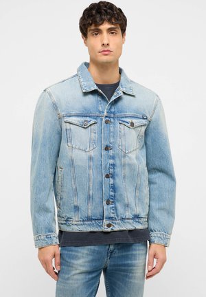 Junger Mann trägt eine hellblaue Jeansjacke und Jeans, steht vor einem schlichten weißen Hintergrund, blickt nach vorne mit neutralem Gesichtsausdruck.