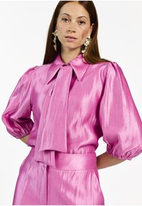 Fuchsia satin blouse with a large collar, puff sleeves, and a knotted tie at the neck. Fabric has a subtle sheen and textured finish.