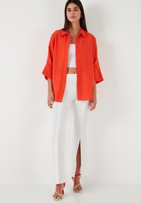 Orange oversized button-up shirt, white cropped tank top, white pants with side slits, and multicolor heeled sandals. Smooth textures.