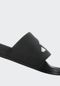 Černá nazouvací pantofle s širokým, texturovaným páskem a lesklým stříbrným logem jako zdobou. Podešev je hladká a mírně tvarovaná pro pohodlí.