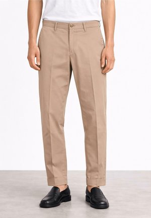 Mężczyzna w beżowych, dopasowanych spodniach z mankietami i czarnych skórzanych wsuwanych butach na białym tle.