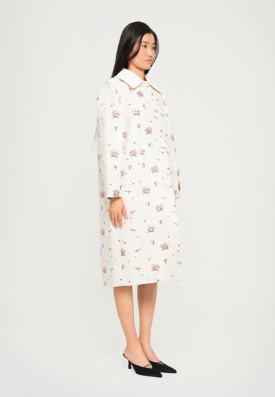 Fehér virágmintás ruha, nagy gallérral és hosszú ujjakkal. Fekete, hegyes orrú magassarkúval viselve. Sima textúrájú, térdhez érő fazon.