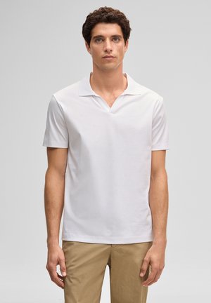 Jonge man met krullend donker haar, gekleed in een wit poloshirt met korte mouwen en beige broek, staand tegen een effen grijze achtergrond.