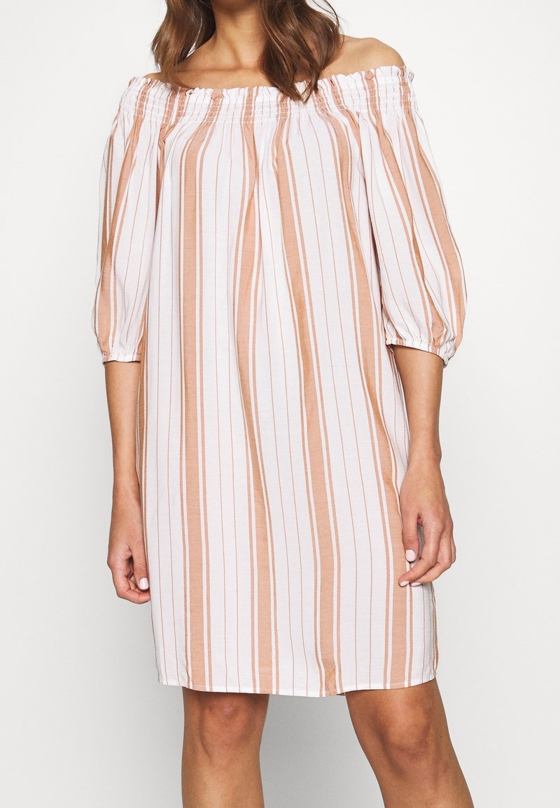 Femme portant une robe longue jusqu'aux genoux, épaules dénudées, à rayures verticales beige et blanches et manches bouffantes trois-quarts.