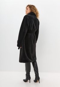Zwarte nepbontjas met een ceintuur in de taille, lange mouwen en knoopdetail bij de manchetten. Draag het met lange zwarte leren laarzen met hoge hakken.