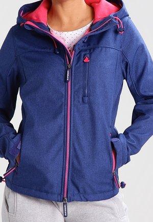 Vrouw draagt een blauw softshelljack met roze rits en capuchonvoering, handen in de zakken, gecombineerd met lichtgrijze broek.