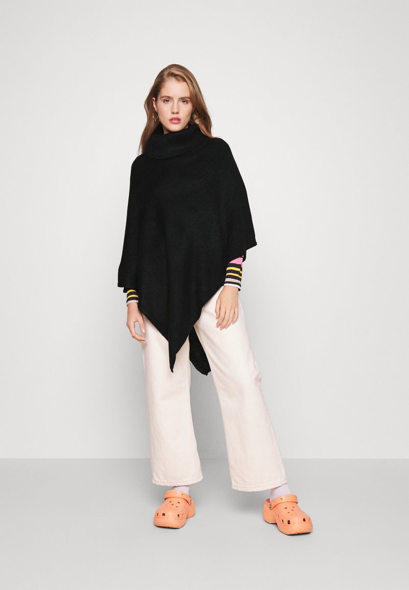 Μαύρο πλεκτό πόντσο με ψηλό γιακά, συνδυασμένο με ανοιχτό ροζ παντελόνια σε ευρεία γραμμή και πορτοκαλί ξύλινες παντόφλες. Ριγέ λεπτομέρειες στα μανίκια.