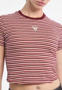 Czerwono-biały w paski crop top z okrągłym dekoltem i krótkimi rękawami. Posiada trójkątną naszywkę logo Guess na piersi. Wykonany z bawełny.