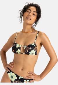 Zwarte bloemenbikini met lichtgroene bloemen, verstelbare dunne straps en een uitgesneden voorkant. Gemaakt van rekbaar materiaal voor comfort.