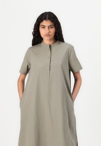 Kratka oblekica z olivno zeleno barvo, izdelana iz mehkega blaga, z gumbi na ovratniku in stranskimi žepi za praktičnost.