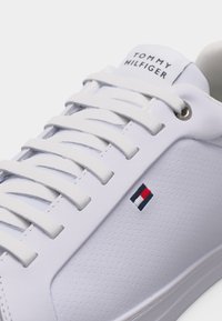 Λευκό δερμάτινο αθλητικό παπούτσι με λευκά κορδόνια, διάτρητο πλαϊνό πάνελ, μικρό κόκκινο, λευκό και ναυτικό λογότυπο, και το κείμενο "Tommy Hilfiger" στη γλώσσα.
