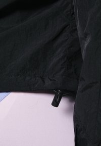 Svart lättviktsjacka med texturerad yta, med dragsko i nederkant och plastspännen, delvis täckande ett ljuslila tyg under.