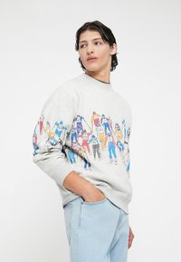 Szary sweter z kolorowymi grafikami w tematyce narciarskiej z przodu, z okrągłym dekoltem i prążkowanymi mankietami. W połączeniu z jasnoniebieskimi dżinsami.