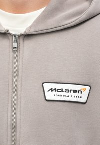 Hallide kapuutsärg, millel on ees lukk. Omab musta ja oranži värvi McLaren Formula 1 Team'i logo, mis on tehtud õmblusega valgel taustal.
