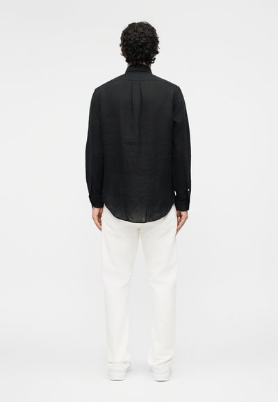 Hombre con cabello rizado, vestido con una camisa negra de manga larga y pantalones blancos, de pie de espaldas contra un fondo blanco liso.