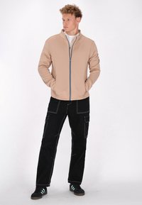 Beige ritsjack met hoge kraag, zwarte cargo-broek met witte stiksels, en zwarte sneakers met teal accenten.