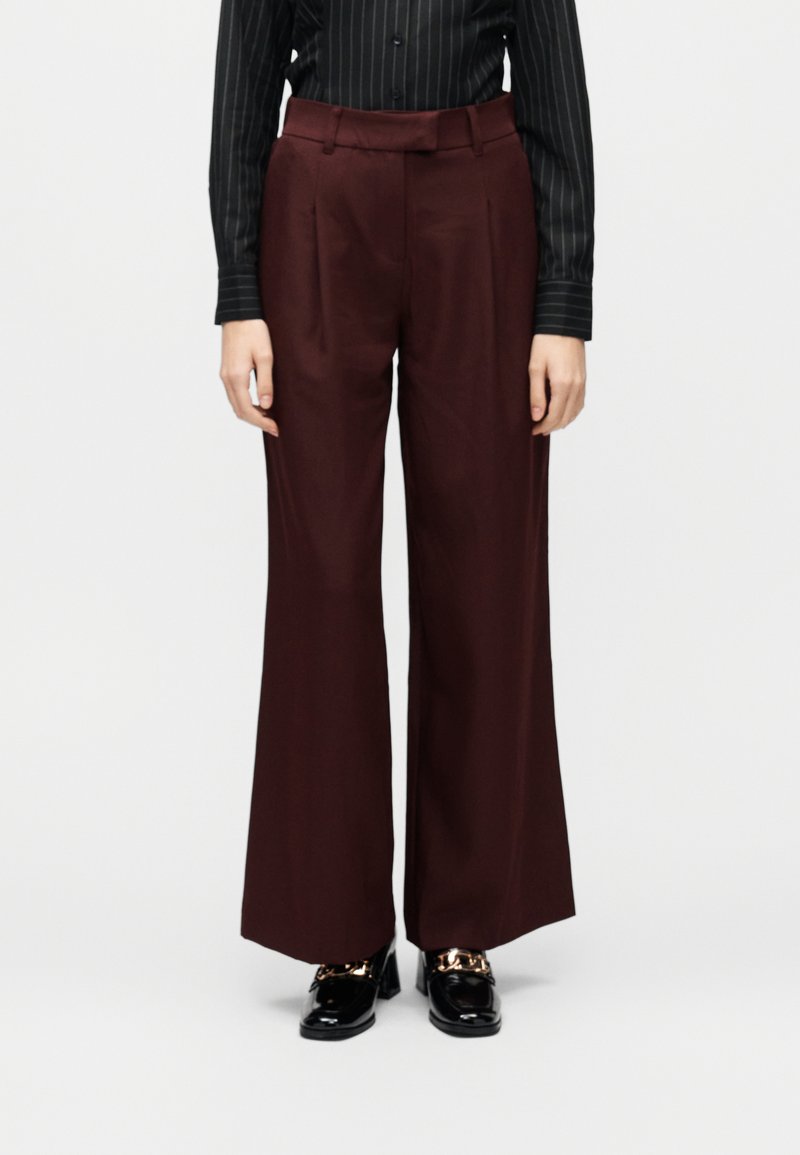 Γυναίκα που φοράει μπορντό παντελόνι ψηλόμεσο με φαρδιά μπατζάκια και κουμπιά μπροστά, με μαύρα παπούτσια loafers από γυαλιστερό δέρμα με χρυσή λεπτομέρεια.