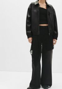 Czarna kurtka z ekoskóry z zapięciem na zamek i elastycznymi mankietami, zestawiona z czarną koronkową narzutą i dżinsami w szerokim kroju, tworząc warstwowe tekstury.