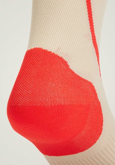 Calcetín beige con un acabado texturizado, que presenta un talón rojo brillante. El diseño incluye una transición de color suave que se fusiona en la zona del talón.