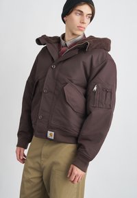 Καφέ μόνωτη ζακέτα με κουκούλα επενδυμένη με ψεύτικη γούνα, κούμπωμα με κουμπιά, μεγάλη μπροστινή τσέπη και τσέπη με φερμουάρ στο μανίκι. Φοράει πάνω από μπεζ παντελόνι.