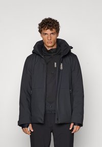 Schwarze gepolsterte Jacke mit hohem Kragen und Kapuze, ausgestattet mit einem Reißverschluss und zwei Fronttaschen. Über einer schwarzen Basisschicht getragen.