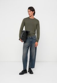 Camisa de manga larga verde oliva, vaqueros rectos de tiro claro en azul con un acabado desgastado y zapatos negros sin cordones, sosteniendo una chaqueta de piel negra.