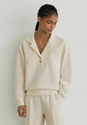 Woman with braided hair wearing a cream long-sleeve shirt with a wide collar and beige pleated pants, standing against a white wall.